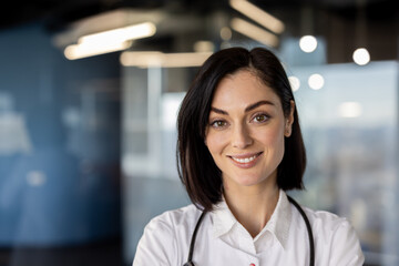 Wall Mural - Confident female doctor with stethoscope smiling at camera in modern medical office. Professional medical staff portrait. Health care and medical services concept.