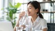 © Vitalii Shkurko - Smiling asian woman drinking water from a bottle while working at a desk with a laptop, surrounded by indoor plants.