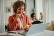 © La Famiglia - Smiling mature businesswoman scrolling a smartphone while working on the computer in the bright office