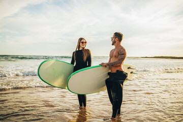 Wall Mural - A couple stands on a sandy beach holding surfboards. They are both wearing wetsuits and sunglasses. The ocean is calm and the sky is clear. The sun is setting in the background