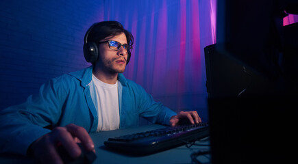 Poster - A man wearing a blue shirt and glasses is focused on his computer screen. He is using a keyboard and mouse, and has headphones on. The room is lit with blue and pink neon lights.