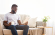 © Prostock-studio - A young black man with a beard sits on a beige sofa in a bright living room, holding a white tablet and a cup of coffee in his hands. He is looking down at the tablet, smiling
