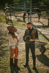 Wall Mural - Young farmer couple with cat in hands standing at ranch with horses.