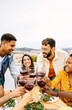 © Xavier Lorenzo - Vertical shot of multiracial group of young friends enjoying bbq dinner party on rooftop. Smiling millennial people celebrating together sitting on terrace table toasting red wine. Copy space.