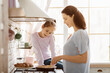 © Prostock-studio - A young girl is learning to cook with her mother in a modern kitchen. The girl is standing at the stove, stirring something in a pan, while her mother watches and smiles