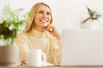 Poster - A woman with long blonde hair smiles while working on her laptop at home. She is sitting at a table with a mug and a plant in the background.