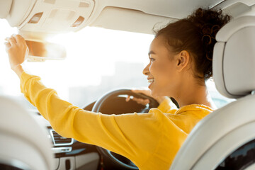 Wall Mural - This image shows a young woman with curly hair sitting in the drivers seat of a car. She is wearing a yellow blouse and smiling as she adjusts the rearview mirror above her head.