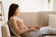 © Prostock-studio - A young woman sits on a beige couch working on a laptop computer.