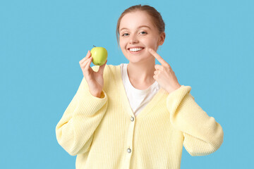 Wall Mural - Young woman with green apple pointing at her teeth on blue background