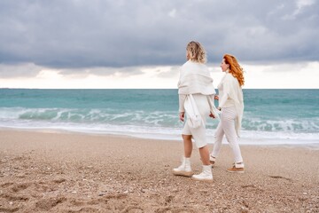 Women sea walk friendship spring. Two girlfriends, redhead and blonde, middle-aged walk along the sandy beach of the sea, dressed in white clothes. Against the backdrop of a cloudy sky and the winter