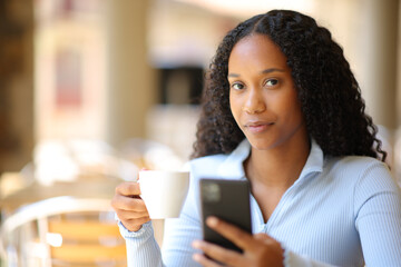 Serious black woman holding phone and coffee in a bar