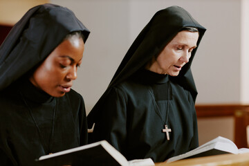 Wall Mural - Medium closeup of two ethnically diverse young and mature Catholic nuns praying while sitting on pew