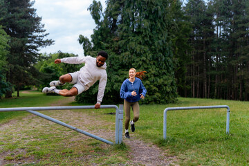 Wall Mural - Man jumping over a barrier while running, with a woman following behind. Both are laughing and having fun during their outdoor workout in the park.