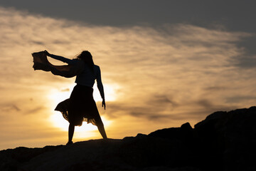 Wall Mural - A woman is standing on a rocky hillside, with the sun setting in the background