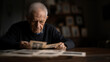 © NaphakStudio - Elderly man looking at old photos in a dimly lit room, dealing with memories and nostalgia.