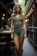 © larrui - Young woman with long blonde hair wearing a green sleeveless outfit with ripped shorts and belt standing in an outdoor urban setting. Street fashion photography. Casual and trendy fashion concept