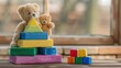© lililia - A colorful stacking toy sits on a white rug in a playroom, with a teddy bear beside a white couch in the background.