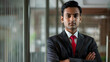 © ramona - an Indian man in his late thirties, wearing a business suit and red tie with blue stripes, standing at the office entrance facing forward to the camera with his arms crossed, smiling softly