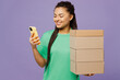 © ViDi Studio - Young smiling happy woman of African American ethnicity with dreadlocks wears green t-shirt casual clothes hold stack cardboard blank boxes use mobile cell phone isolated on plain purple background.