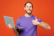 © ViDi Studio - Young smiling cheerful fun happy IT man he wearing purple t-shirt casual clothes hold use work on laptop pc computer show thumb up isolated on red orange background studio portrait. Lifestyle concept.