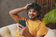 © ViDi Studio - Young Indian man wear orange casual clothes hold head use mobile cell phone read news sits in armchair stay at home hotel flat rest relax spend free spare time in living room indoor. Lounge concept.
