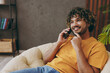 © ViDi Studio - Young cheerful fun Indian man wear orange casual clothes talk speak on mobile cell phone sits in armchair stay at home hotel flat rest relax spend free spare time in living room indoor Lounge concept