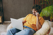 © ViDi Studio - Young IT man in orange casual clothes use work on laptop pc computer talk on mobile cell phone sits in armchair stay at home hotel flat rest relax spend free spare time in living room Lounge concept