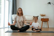 © standret - Calm, in lotus pose. Young woman with little girl are doing yoga at home