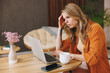 © ViDi Studio - Young sad tired confused IT woman she wears orange shirt hold use laptop pc computer sit alone at table in coffee shop cafe restaurant indoor work or study. Freelance mobile office business concept.