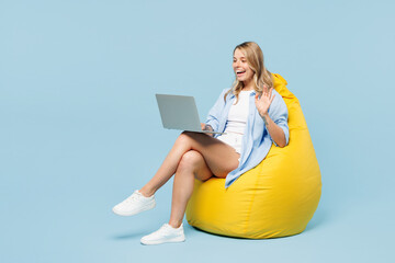 Wall Mural - Full body young happy woman she wears white top shirt casual clothes sit in bag chair hold use work on laptop pc computer waving hand isolated on plain pastel light blue background. Lifestyle concept.