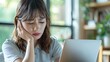 © naphat - A Japanese woman is sitting at her desk in the living room, holding her neck in pain and using a laptop computer for work. She has shoulder-length hair with bangs on one side of her head, wearing