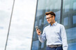 © muse studio - A smiling young professional in a light blue shirt, standing against a modern glass building, using a smartphone.