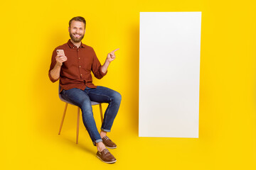 Full size photo of positive guy sit chair direct finger empty space telephone screen isolated on yellow color background