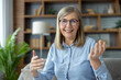 © Liubomir - Senior woman with glasses engaging in a video call while looking and talking to the camera. She seated in a cozy living room with shelves and plants in the background, smiling warmly and gesturing.