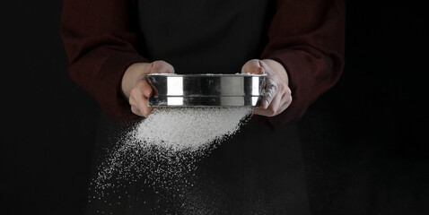 Wall Mural - Woman sieving flour at table against black background, closeup