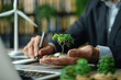 © btiger - Businessman Holding Coins with Tree Growing, Laptop, and Wind Turbine in Office for Green Energy Concept. Investment Ideas, Sustainable Finance, Eco Business Growth, Alternative Energy Sources