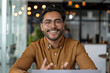 © btiger - Confident latin man in casual wear and glasses sitting at desk in modern office smiling and talking to camera during video call or remote training session