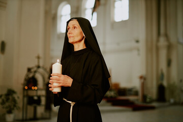 Wall Mural - Medium shot of mature Caucasian Catholic nun holding burning candle in her hands standing inside church building