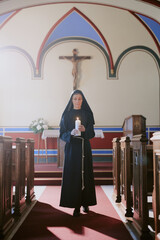 Wall Mural - Vertical wide shot of senior Caucasian nun in black habit holding burning candle walking along nave in Catholic church