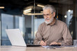 © Liubomir - Senior businessman working on a laptop and taking notes in a modern office. The image captures a mature professional in a work environment, showcasing productivity, experience, and dedication