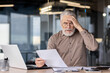 © Liubomir - Senior businessman looking stressed while reading documents at his office desk. He is sitting in front of a laptop, holding a paper and appears to be in deep thought or confusion.