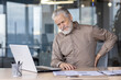 © Liubomir - Senior man suffering from back pain while sitting at his office desk and working on a laptop. This image captures discomfort, health, and ergonomic concerns in a professional office environment.