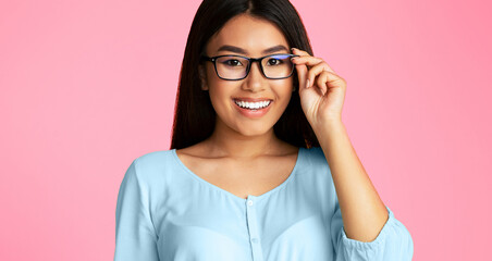 Asian young woman with long black hair is smiling while adjusting her black framed glasses. She is wearing a light blue shirt and is standing against a pink background.