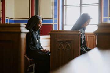 Wall Mural - Side view shot of two diverse nuns sitting on wooden benches in Catholic cathedral or church praying to Lord