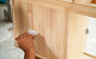 Carpenter hands applying varnish to a cabinet with a paintbrush. First lacquer coating on wood surface at a furniture and wooden workshop.Close up.