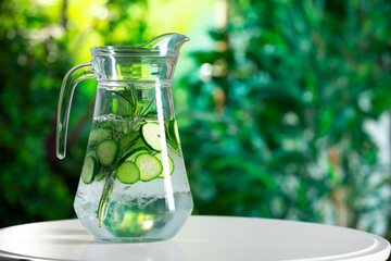 Refreshing cucumber water with rosemary in jug on white table against blurred green background. Space for text