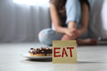 Wall Mural - Eating disorder. Woman sitting on floor indoors, focus on sticky note with word Eat and donut