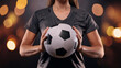 © desire - Igualdad futbol femenino. Mujer deportista irreconocible sujeta un balón de fútbol entre sus manos con fondo oscuro y desenfocado de fondo. La atleta lleva una camiseta deportiva negra.