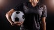 © desire - Fútbol femenino. Mujer futbolista con camiseta negra de deporte sujetando con una mano un balón de fútbol. Concepto de igualdad, superación empoderamiento