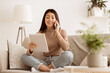 © Prostock-studio - A young woman sits on a couch in a living room, talking on her phone while looking at a tablet. She is wearing a beige sweater and blue jeans.